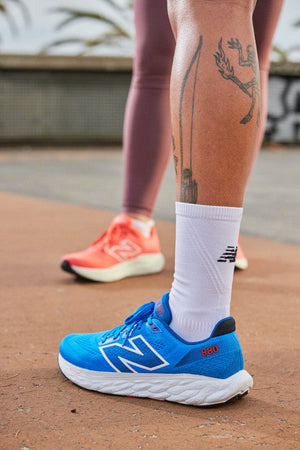 Two runners' legs wearing New Balance shoes. The runner's leg in the foreground is a back view and he has tattoos on the calf, white socks and blue shoes. The runner in the background is wearing orange running shoes.
