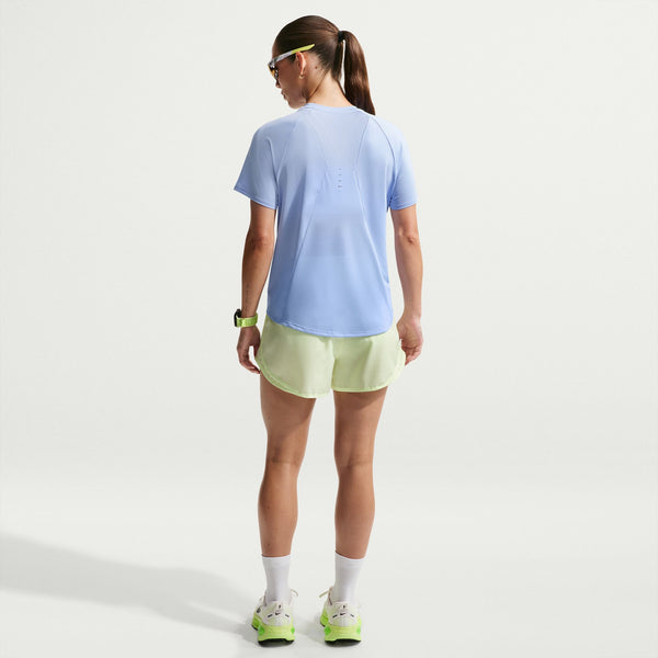 A woman is shown from behind wearing a light blue athletic t-shirt, lime green running shorts, white socks, and neon green running shoes against a plain white studio background.