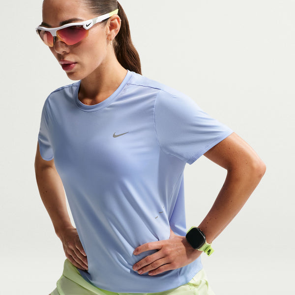 A woman wearing a light blue Nike athletic short-sleeve t-shirt, white running sunglasses with pink lenses, and a smartwatch, posed against a light gray studio background.