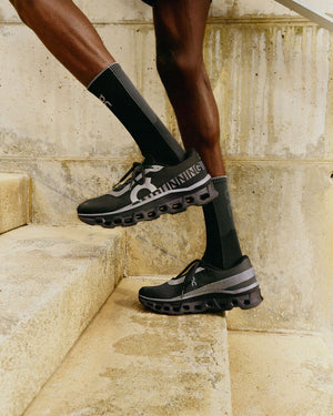 A left lateral view of a male runner's legs from the knees down in black On Cloudmonster shoes and black socks going up steps with a warm neutral color tone for the concrete