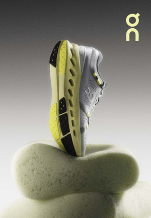 Image of the new Cloudsurfer 2 by ON in a vertical position with the toe down sitting on a piece of foam to showcase the pillowy nature of the midsole. Color scheme of photo is grey and yellow hues. The ON logo is displayed in the top right corner in yellow.