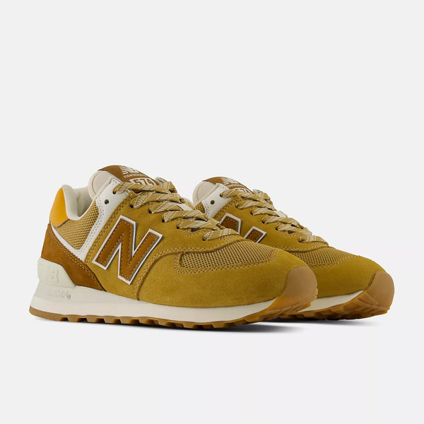 A pair of New Balance 574 sneakers in a mustard yellow and brown colorway, featuring a white 'N' logo and white midsoles, set against a light gray background.
