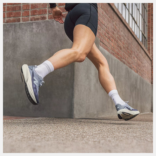 A person running outdoors wearing black athletic shorts, white socks, and New Balance running shoes in navy, white, and gray, with a brick and concrete wall in the background.