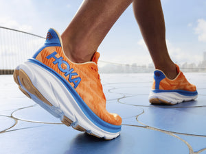 Up close view of the right foot of a male taking a step with the heel coming up wearing Bright orange and blue Hoka Clifton shoes on circular pavers and a blurry distance background of the sky and clouds.