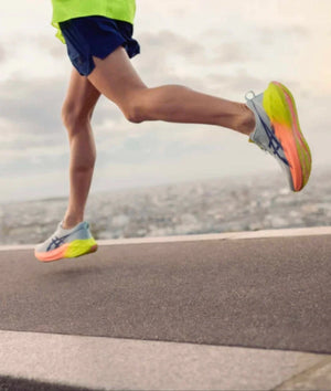 A view of a male runner from the back and waist down wearing Brightly colored yellow and pink ASICS running shoes, navy shorts and a yellow shirt with a neutral blurry background