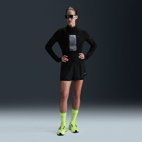 A model wearing a black Nike running jacket, black running shorts, neon yellow socks, and neon yellow running shoes, posing against a dark gray studio background.
