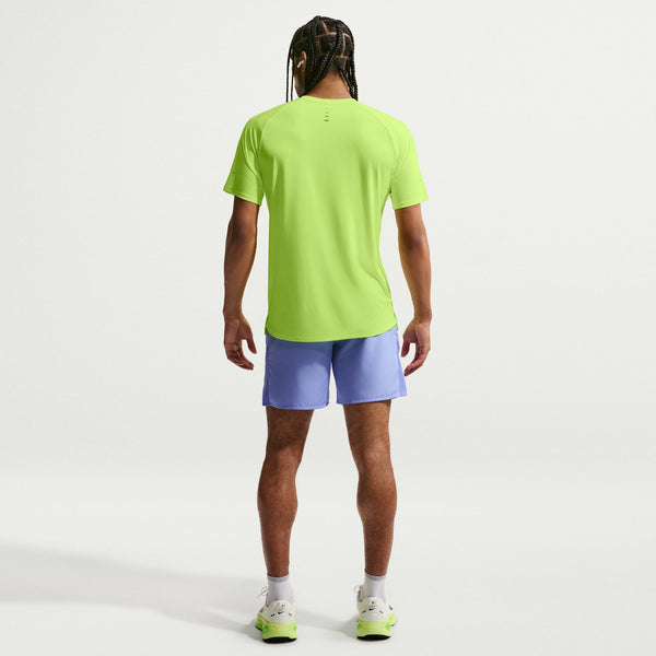 A man viewed from behind wearing a bright lime green athletic t-shirt and light purple athletic shorts, standing against a clean white studio background.