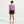 A woman viewed from behind wearing a purple athletic t-shirt, black athletic shorts, white socks, and pink and black running shoes against a light gray studio background.