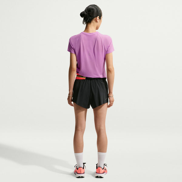 A woman viewed from behind wearing a purple athletic t-shirt, black athletic shorts, white socks, and pink and black running shoes against a light gray studio background.