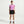 A woman stands against a light gray studio background wearing a light purple short-sleeve athletic T-shirt, black athletic shorts, white socks, and pink and black running shoes.