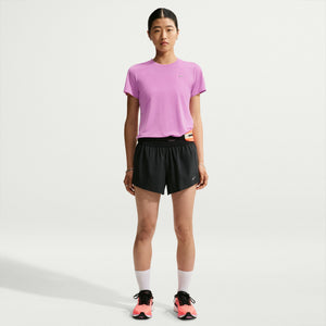 A woman stands against a light gray studio background wearing a light purple short-sleeve athletic T-shirt, black athletic shorts, white socks, and pink and black running shoes.