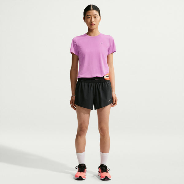 A woman stands against a light gray studio background wearing a light purple short-sleeve athletic T-shirt, black athletic shorts, white socks, and pink and black running shoes.