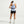 A woman is shown from behind wearing a light blue athletic tank top, black athletic shorts, white socks, and white sneakers against a plain light gray studio background.