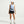 A woman modeling athletic wear in a studio setting, wearing a light blue sleeveless top, dark navy shorts, a black waist belt, a white cap, white socks, and white sneakers.