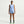 A model stands against a light gray studio background wearing a light blue sleeveless athletic tank top and matching light blue athletic shorts with dark trim, black socks, and colorful running shoes.