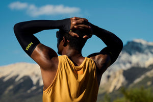 The back view of a male with his hands linked and resting on the back side of his head.  The image is from the waist up and his tank top is orange.  The background is a blurry view of a mountain and blue sky.