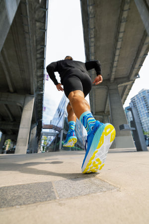 A close up of male runner's shoes running away from you.  The bottom soles are visible and are white and yellow. The runner is in an urban scene wearing a black long sleeve and black short tights.