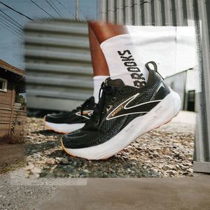 Up close lateral view of the left foot of a male wearing Black Brooks running shoes and white socks with the word Brooks in black on the socks.  The background is a blurred out gritty urban scene.