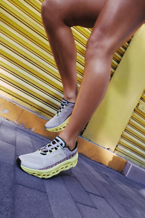 Photo of a female's legs from the thigh down emphasizing her running shoes.  She is tan with grey and yellow shoes in front of a yellow wall.