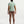 A woman viewed from behind wearing a light sage green athletic short-sleeve t-shirt and mauve-brown leggings, standing against a clean white studio background.