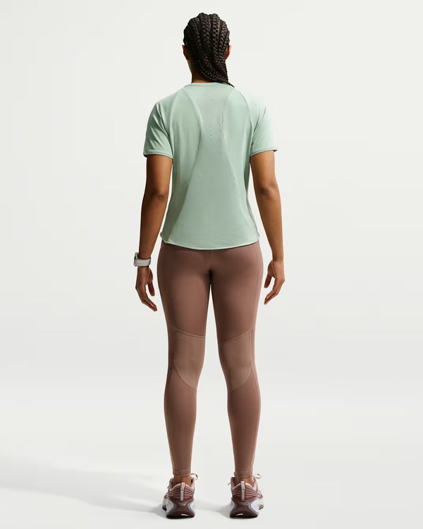 A woman viewed from behind wearing a light sage green athletic short-sleeve t-shirt and mauve-brown leggings, standing against a clean white studio background.