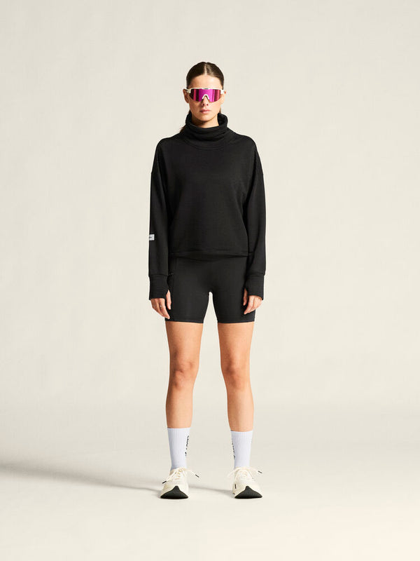 Person wearing a black turtleneck, black shorts, white socks, and black sneakers on a beige background