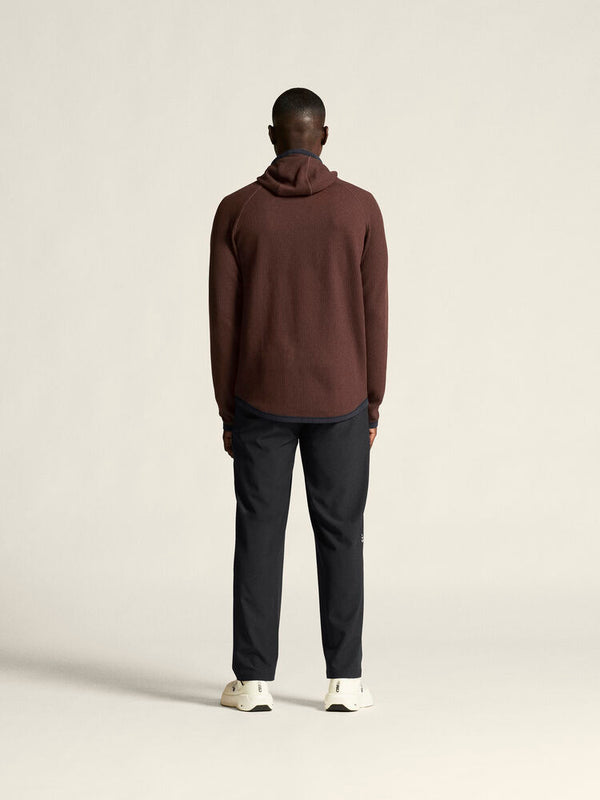 Person wearing a brown hoodie and dark pants on a beige background