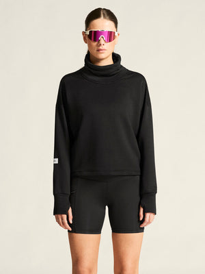 Person wearing a black sweatshirt and shorts with reflective sunglasses on a beige background