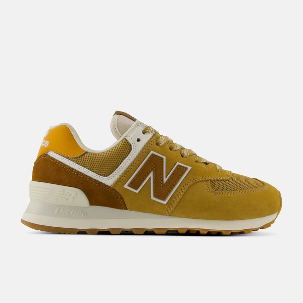 A New Balance 574 sneaker in a mustard yellow and brown colorway, featuring a cream midsole and white trim, shown from a side profile on a light gray background.