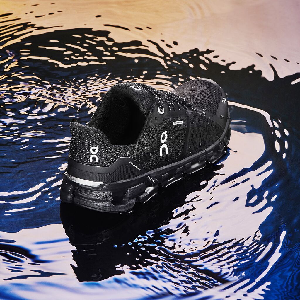 Back angled view of the Women's Cloudflyer -Waterproof by On in the color Black / Lunar on a puddle of water